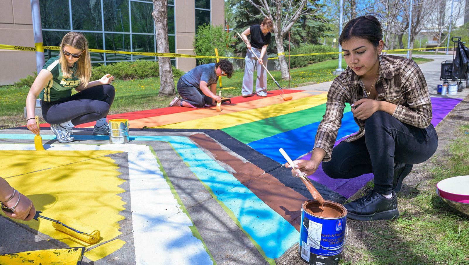 Several students paint different sections of a rainbow crosswalk on a campus sidewalk, using brushes and rollers with paint supplies placed nearby.