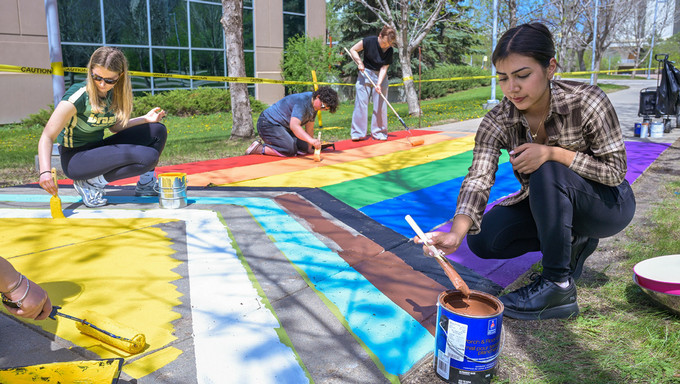 Several students paint different sections of a rainbow crosswalk on a campus sidewalk, using brushes and rollers with paint supplies placed nearby.