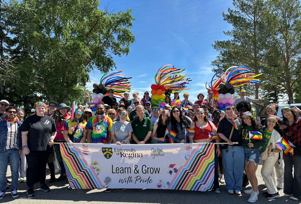 Large group of University of Regina participants posing behind a banner reading “Learn & Grow with Pride,” with rainbow balloon decorations above.