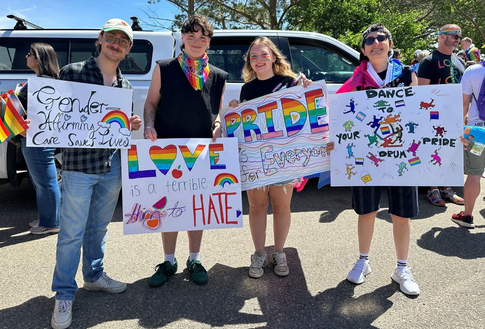 Four parade participants standing side by side holding hand-drawn Pride signs with messages supporting inclusion, gender-affirming care, and community