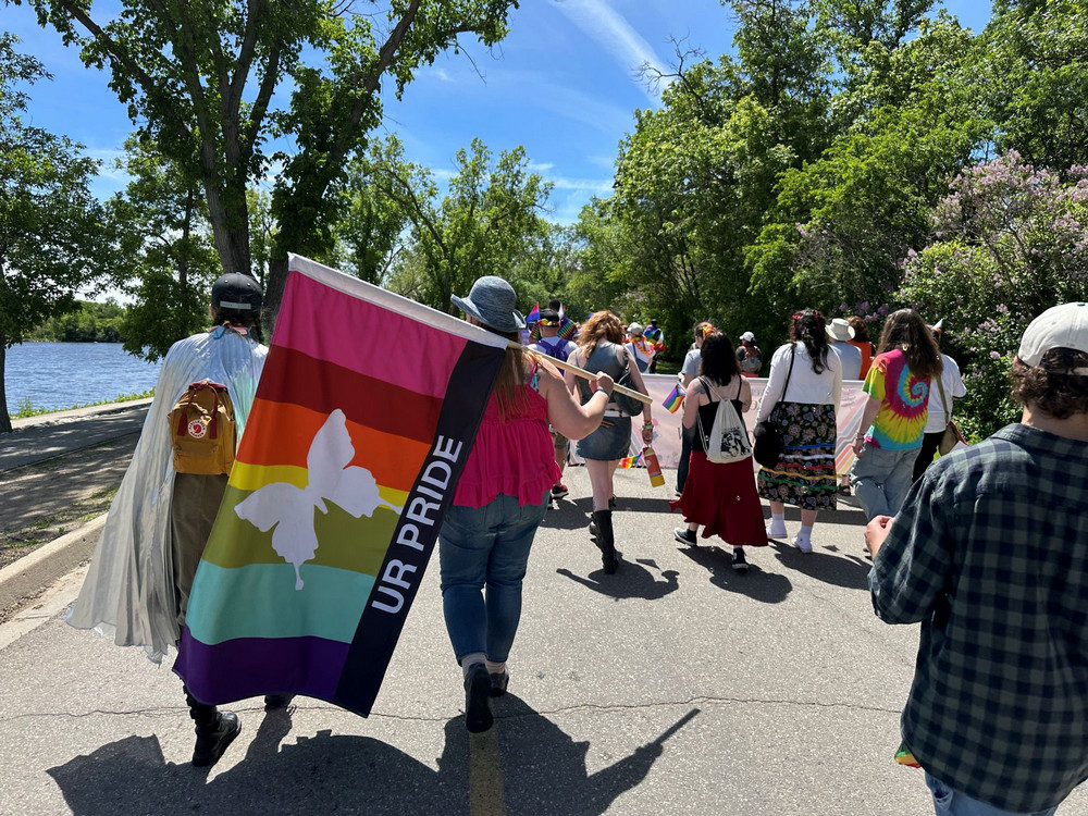 Parade participants walking along a path, one carrying a large rainbow flag with a white butterfly emblem and “UR Pride” text