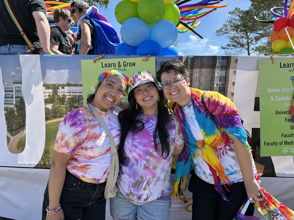Three parade participants wearing tie-dye shirts stand together in front of a decorated float with rainbow balloons