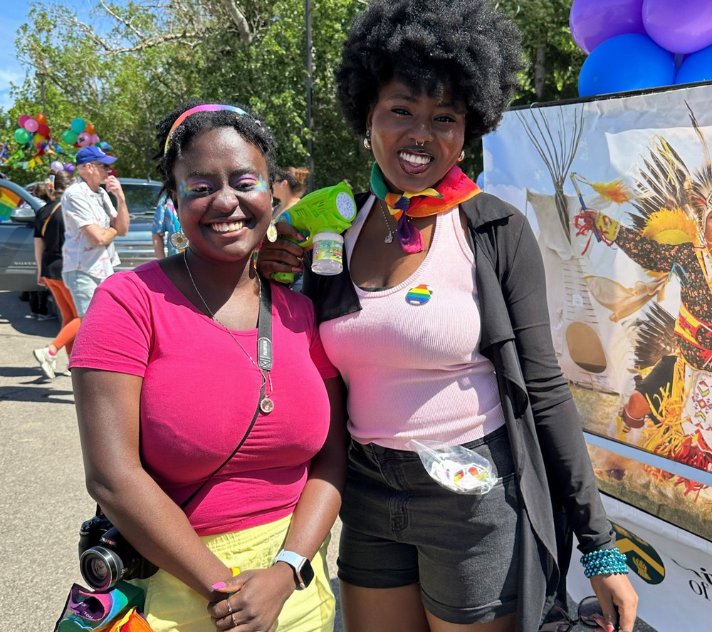 Two parade participants standing together, one holding a green bubble gun, both wearing bright clothing, with a Pride float behind them