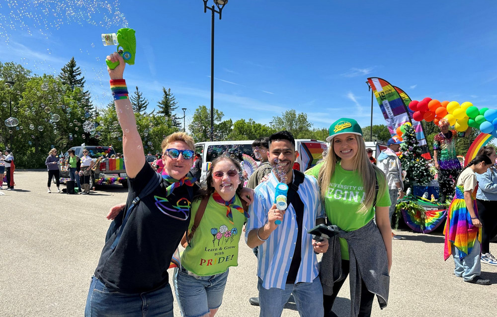 : Four University of Regina participants stand together at a Pride parade, one raising a green bubble gun as bubbles float overhead, with decorated vehicles and a rainbow balloon display in the background.
