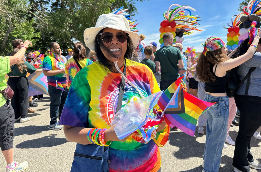 University of Regina parade participant wearing a rainbow tie-dye shirt and white hat holds small Pride flags while standing among a crowd near a decorated float.