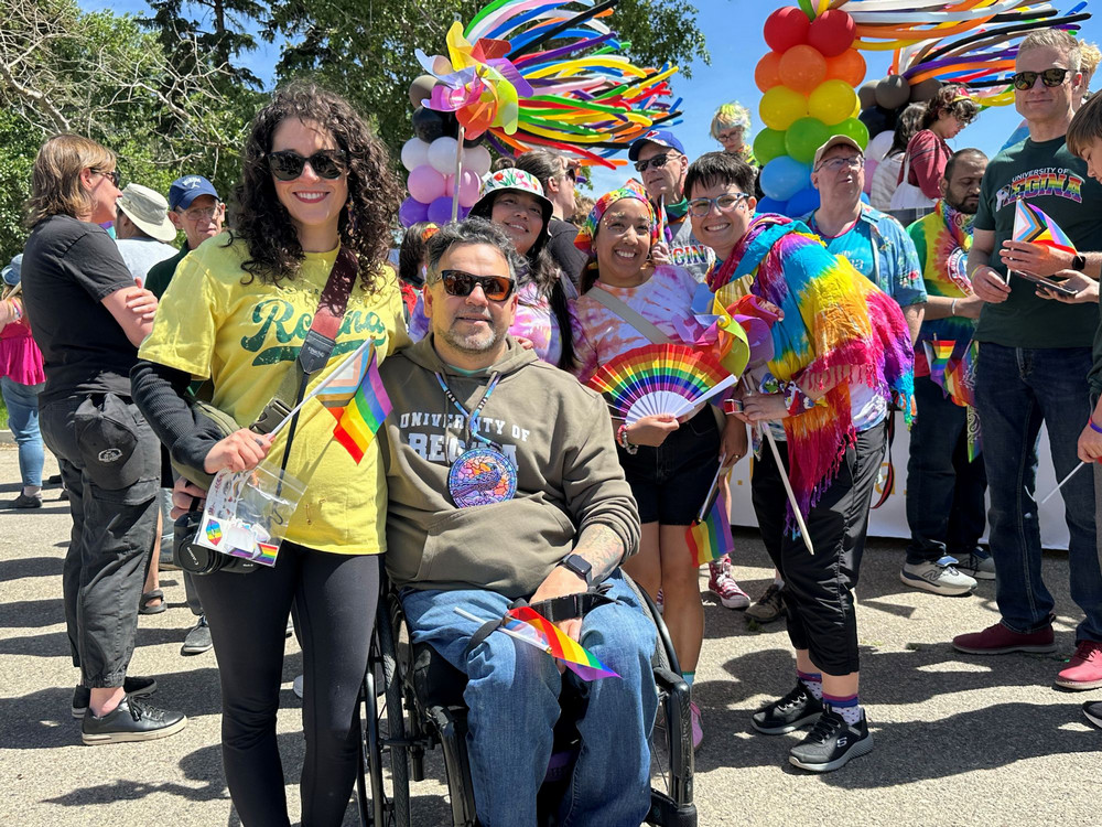 A group of University of Regina participants gathered at a Pride parade, including a person seated in a wheelchair holding a small rainbow flag, surrounded by others holding flags.
