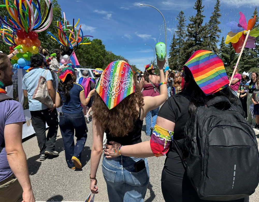 Parade participants walking down a city street behind a decorated float, holding rainbow flags and pinwheels, with trees and vehicles along the route
