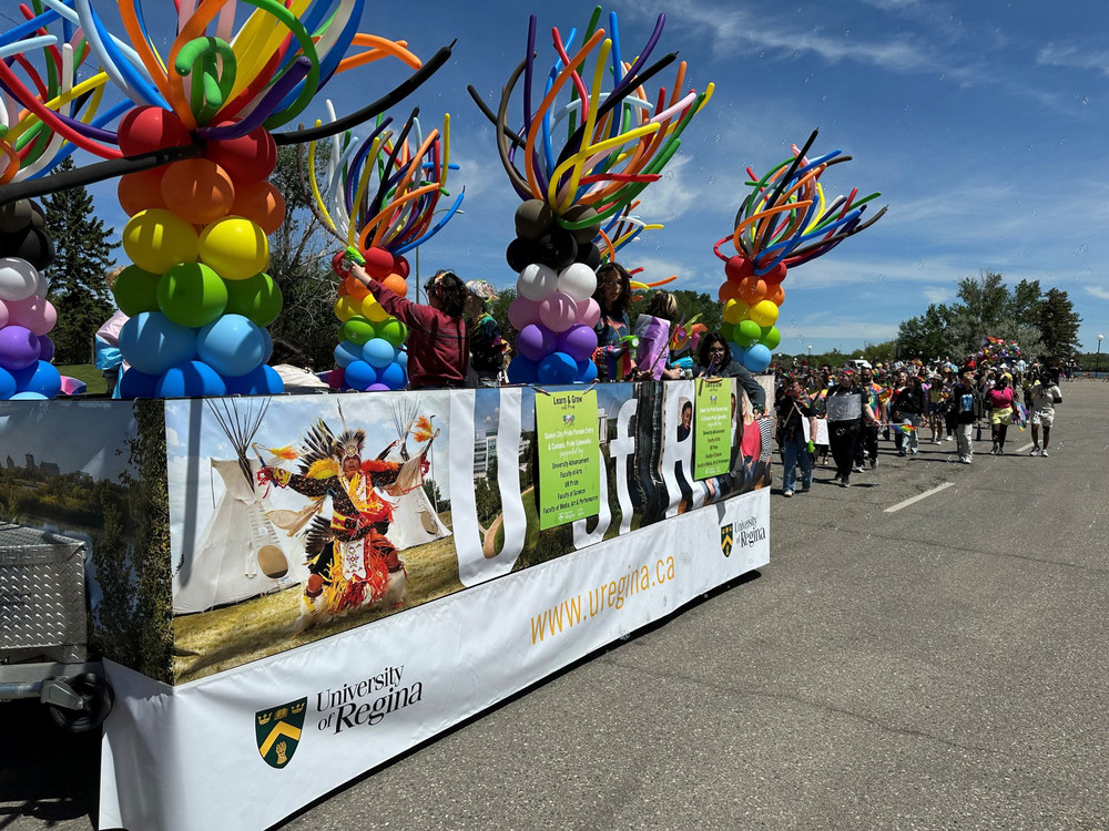 University of Regina Pride float decorated with rainbow balloon columns moves along the parade route as participants walk beside it.