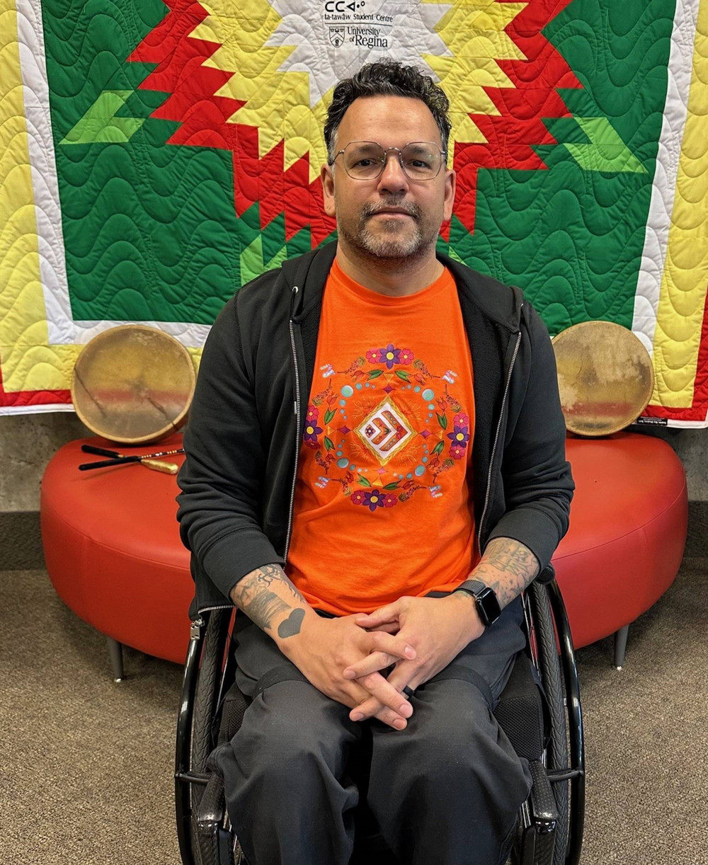 A man poses in front of a colourful blanket on the wall behind him