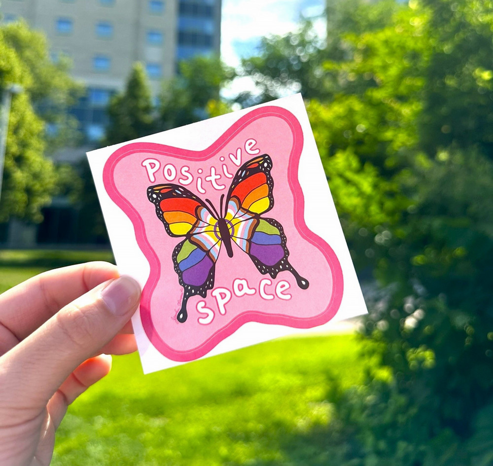 A hand holds a Positive Space sticker featuring a rainbow-coloured butterfly and the words “Positive Space” against a blurred outdoor campus background with trees, grass, and a building.