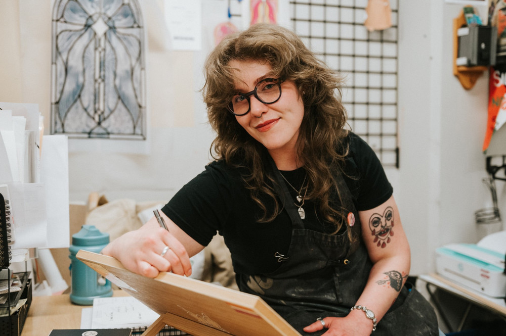 A person seated in an art studio holds a drawing board while surrounded by art supplies, sketches, and creative workspace materials.