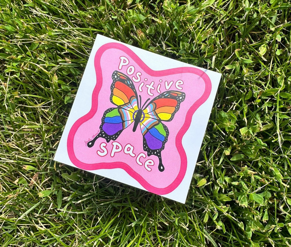 A Positive Space sticker featuring a rainbow-coloured butterfly and the words “Positive Space” rests on green grass outdoors.