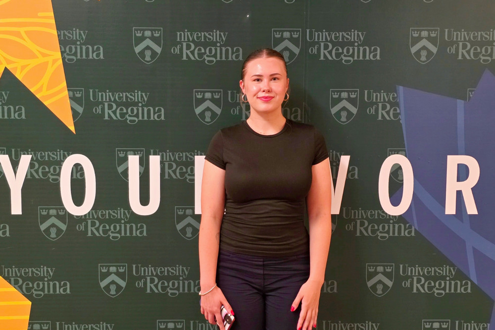 Shantelle Loke, a 4th year Psychology student in the Faculty of Arts, stands in front of a University of Regina-branded backdrop.