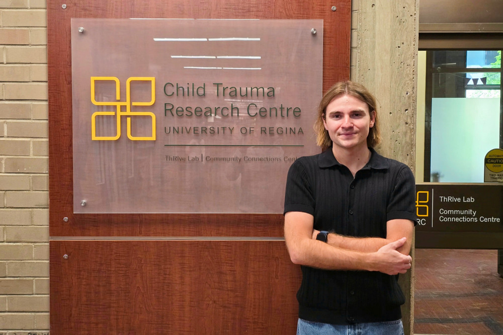 Gavin Gilhooly, a 4th year Psychology student in the Faculty of Arts, stands with arms crossed beside the Child Trauma Research Centre sign at the University of Regina, where he completed his AWEI research placement.
