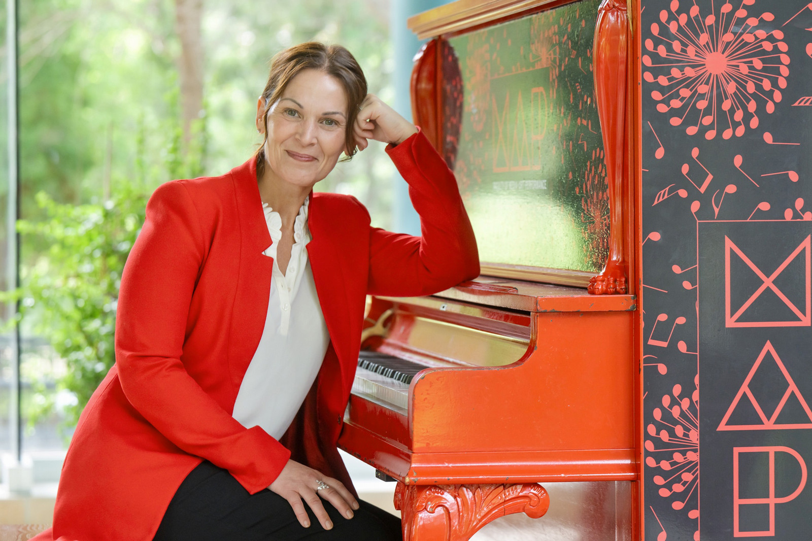 A woman in a very red blazer sits at a piano stool of a red piano.