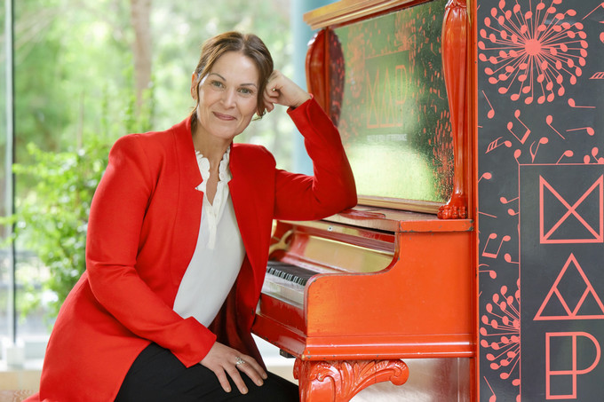A woman in a very red blazer sits at a piano stool of a red piano.