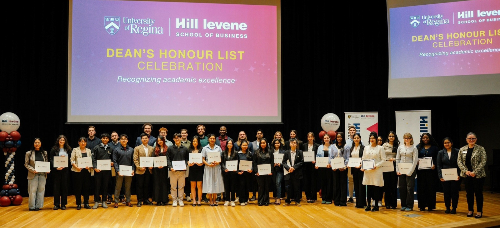 Hill Levene students and employees stand across the Education Auditorium stage holding Dean's Honour List certificates.