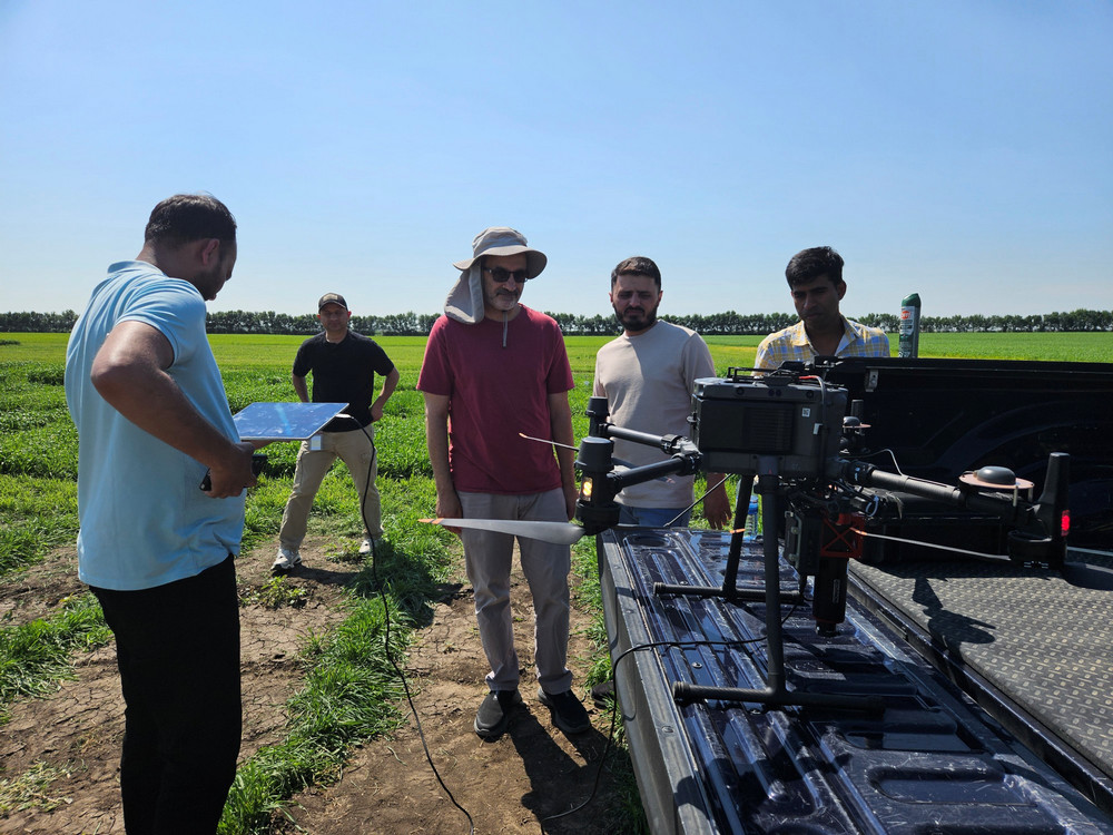 Five people gather at the open tailgate of a pickup truck at the edge of a green field. A large multi-rotor drone and its sensor payload are mounted on the tailgate, and one person holds a tablet.