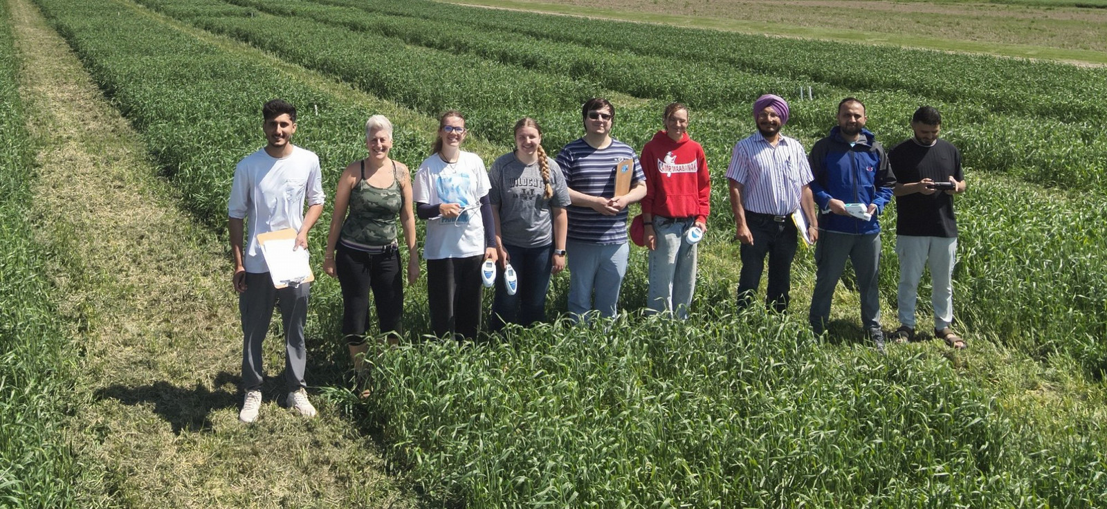 A group of nine people stands side-by-side, smiling in a green agricultural field. Farm buildings and grain bins are visible in the background under a blue sky.
