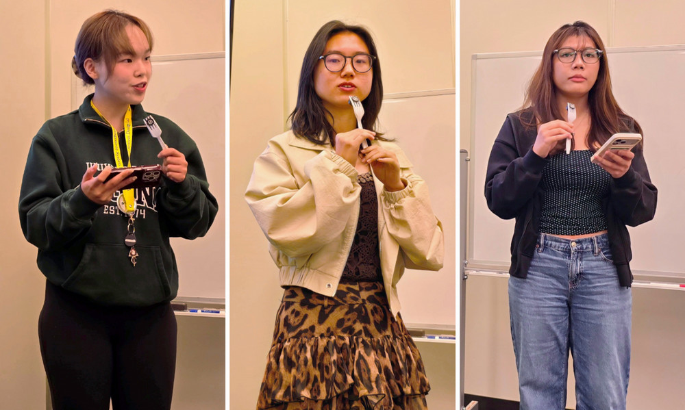 Three students stand side-by-side during an informal presentation, speaking to an audience while holding a microphone.