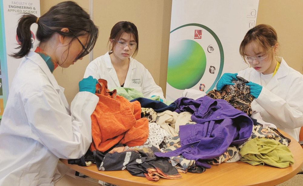 Three students in white University of Regina lab coats and blue gloves examine donated clothing spread across a table 