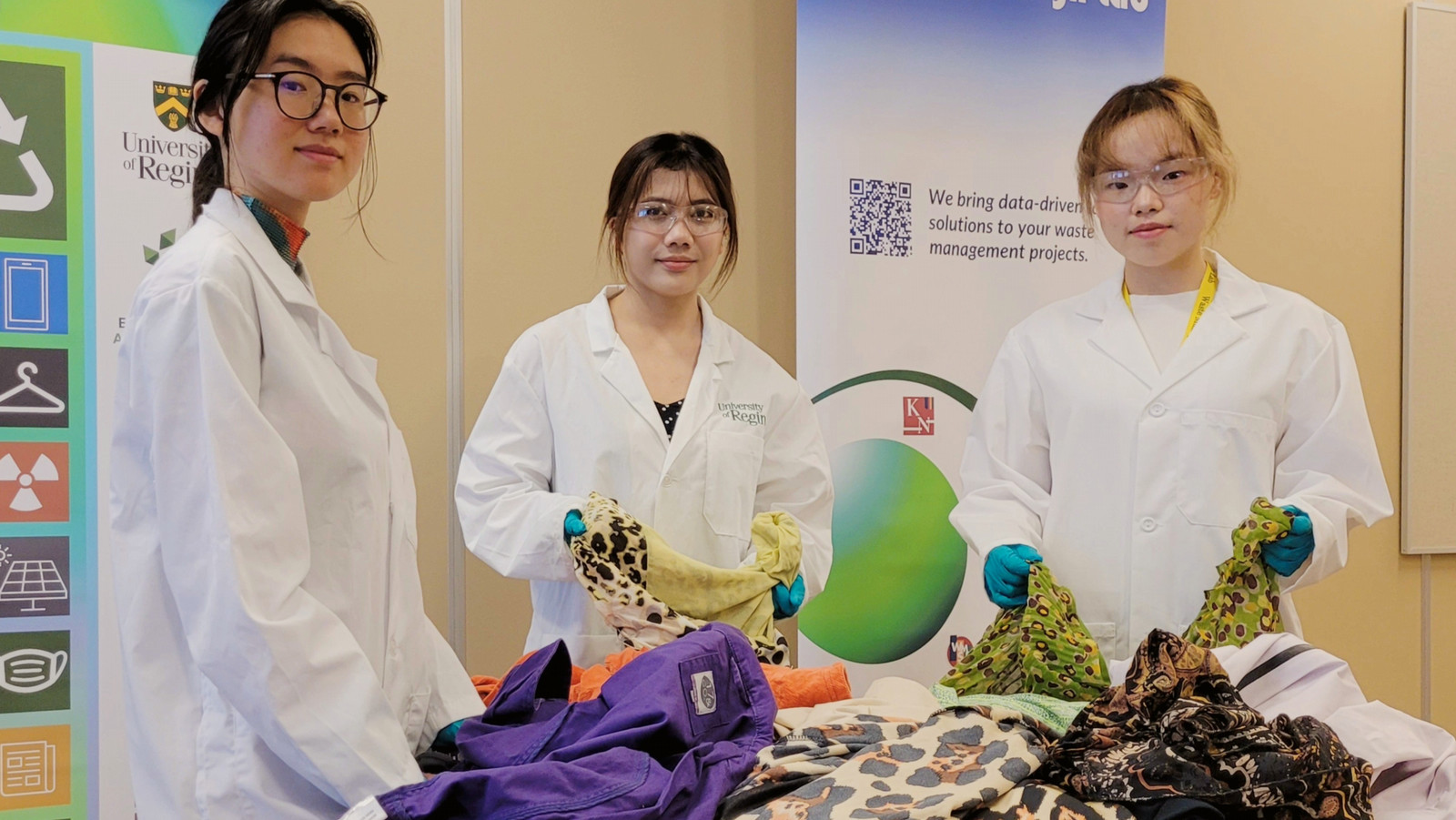 Three students in lab coats and protective eyewear look toward the camera from behind a table covered with sorted second-hand clothing.