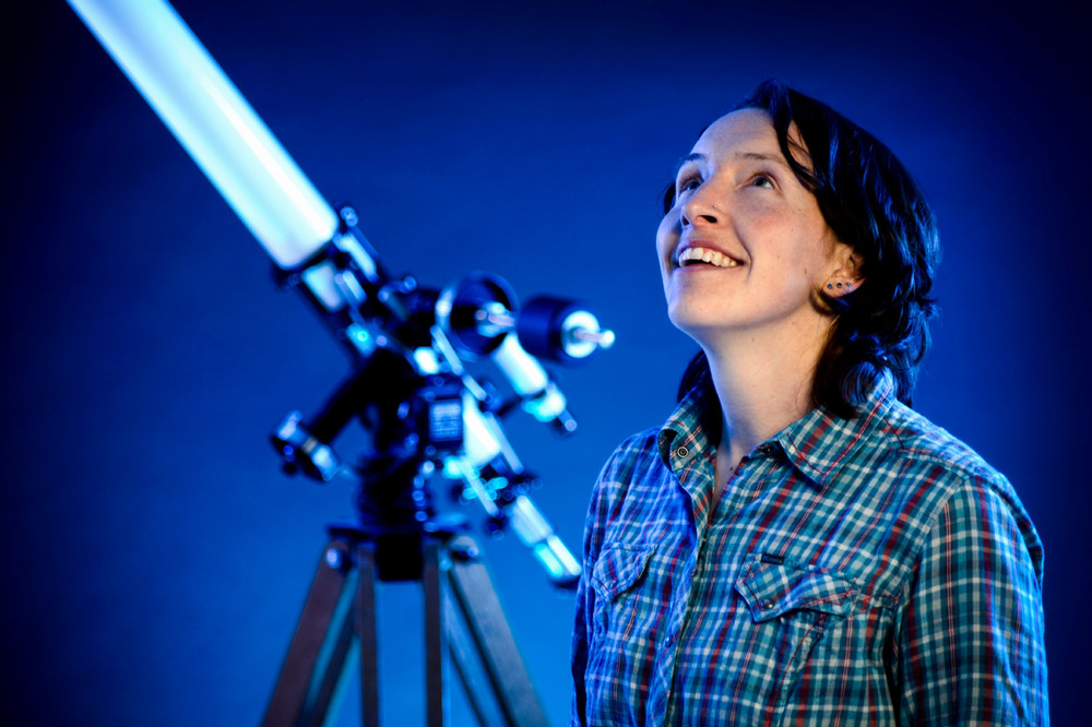A young woman gazes upwards with a telescope in the background