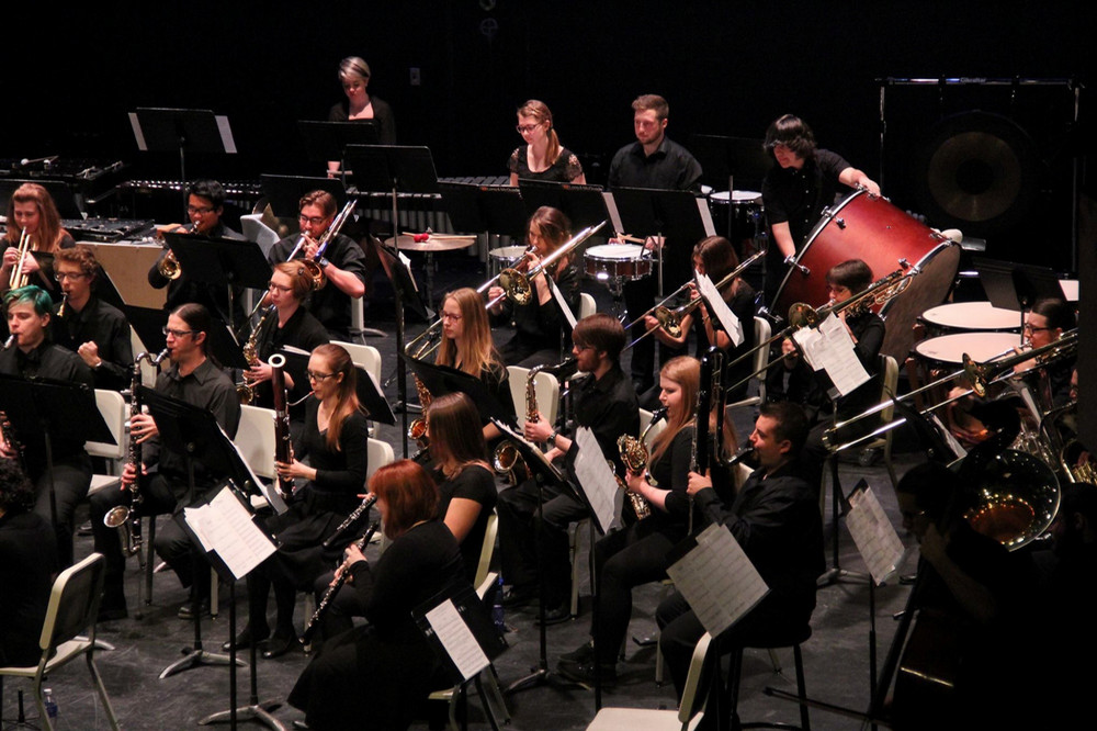 A large ensemble of University of Regina musicians performs on stage, with students playing brass, woodwind, and percussion instruments during a concert.