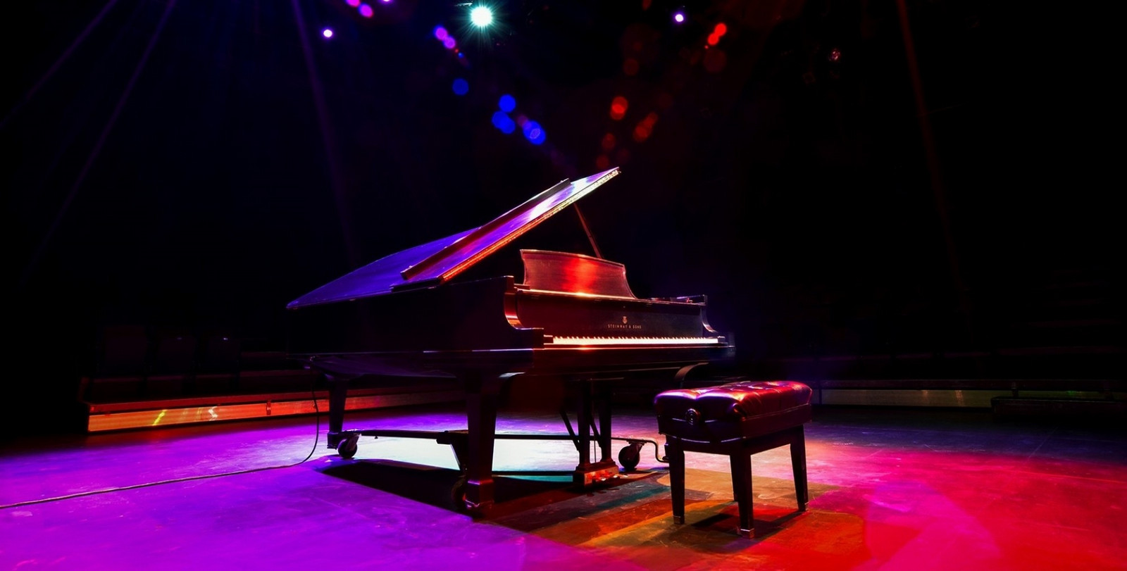 A grand piano sits on the stage of the University of Regina's Riddell Theatre under colourful theatrical lighting.