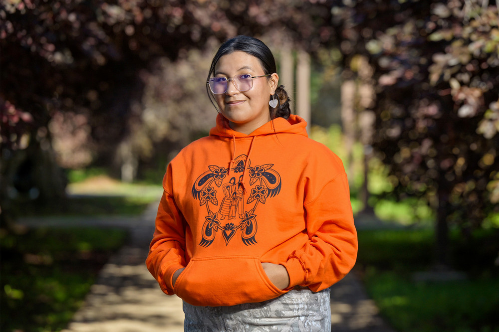 A person wears an orange hoodie featuring her winning Orange Shirt Day design.