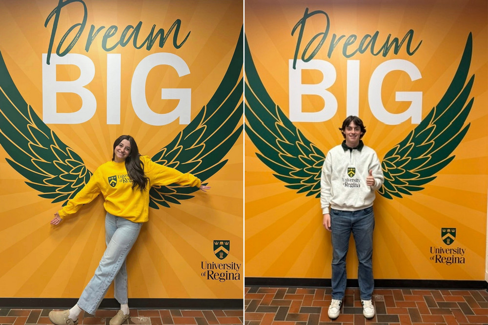 two students pose in front of a yellow Dream Big mural with green wings 