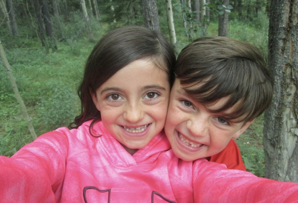 two young children smile closely together outdoors in a forest.
