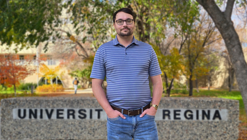 a man stands in front of a University of Regina sign on campus.