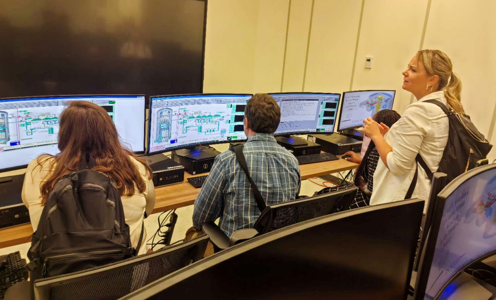 Three people sit at a row of computer monitors showing reactor system diagrams while a woman stands beside them, watching one of the screens.