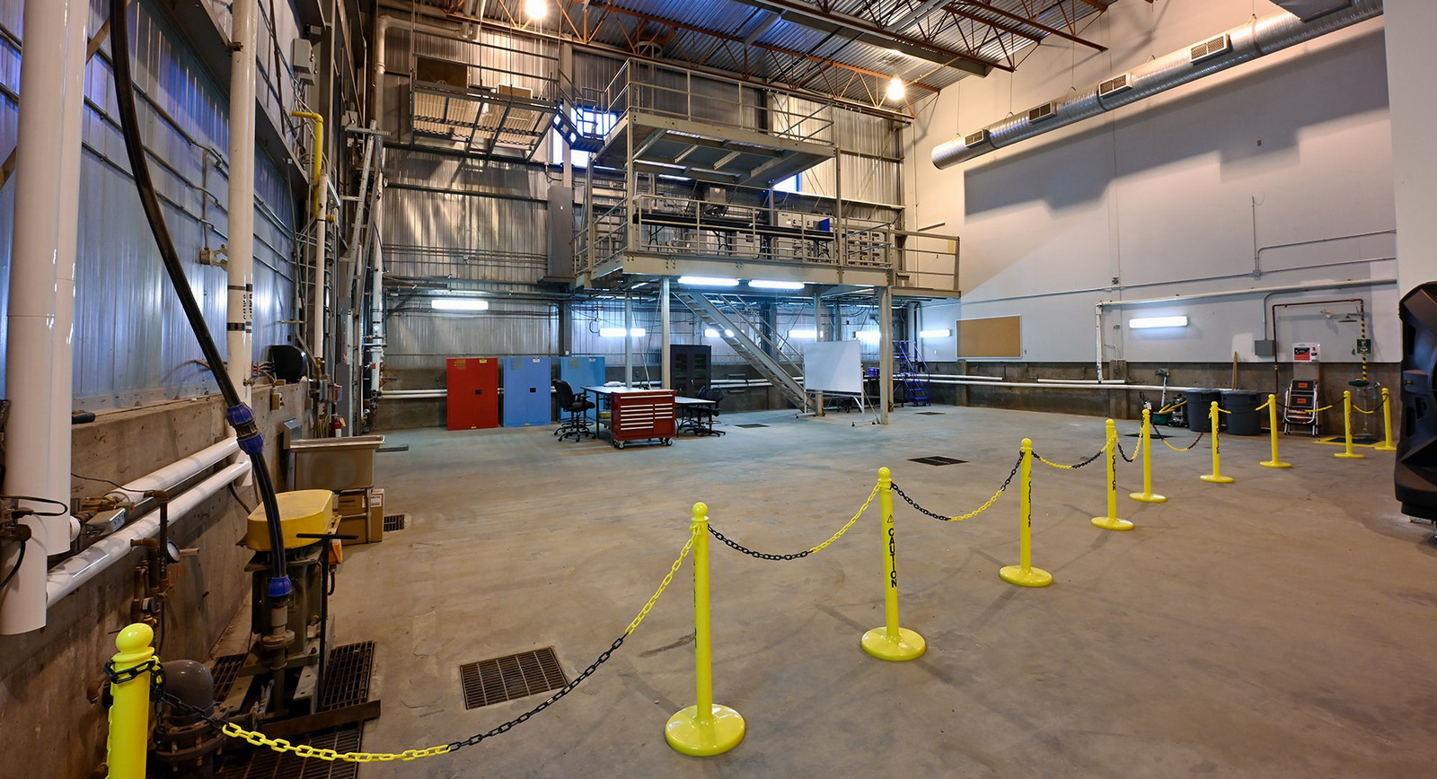 A high-ceilinged hall with a steel mezzanine, ductwork, and yellow barrier posts joined by chain.