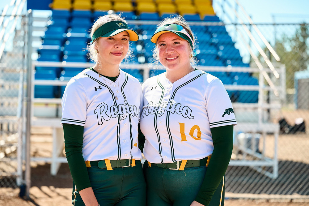 Two young women in baseball uniforms pose in front of bleachers at a ballpark.