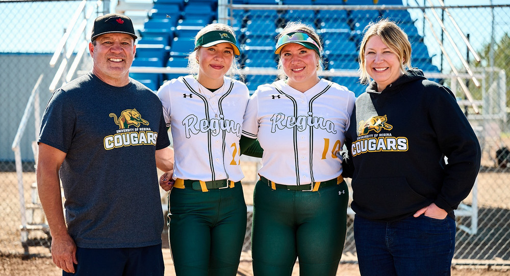 A family of four pose in front of bleachers at a baseball field. 
