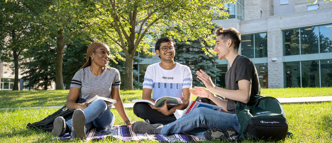 Three students studying and chatting on a lawn in front of a campus building.